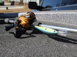 The Lew's Custom Pro Gen 3 casting reel is laying on a sunny bass boat deck with a jerkbait attached to its integrated Speed Keeper bait keeper.